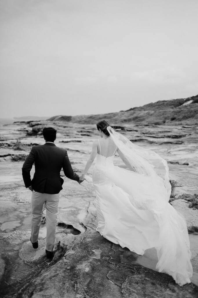 Black-and-white photo of Jeannette and Pierre walking hand in hand across the rock platform at Cape Banks, La Perouse, her veil streaming behind.