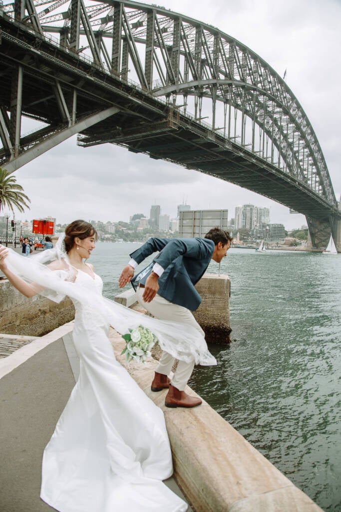 Jeannette holds her veil as Pierre leans over a stone wall near Pylon Lookout beneath Sydney Harbour Bridge.