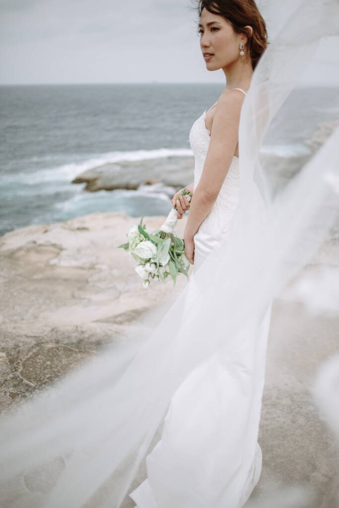 Jeannette stands in profile with her bouquet on a Cape Banks rock platform, her veil crossing the foreground and surf behind.