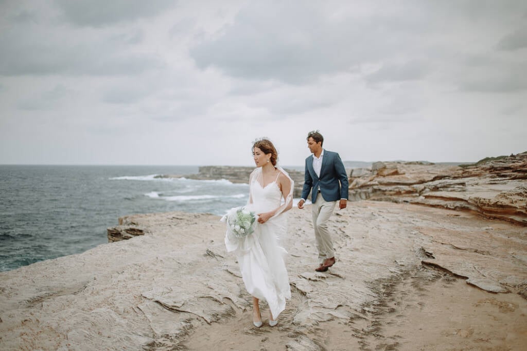 Jeannette walks ahead of Pierre across the sandstone at Cape Banks under a cloudy sky.