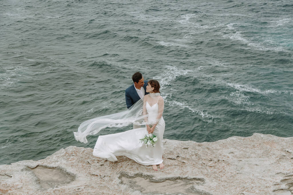 At Cape Banks, Jeannette stands barefoot on a sandstone ledge with Pierre behind her as her veil blows over the sea.