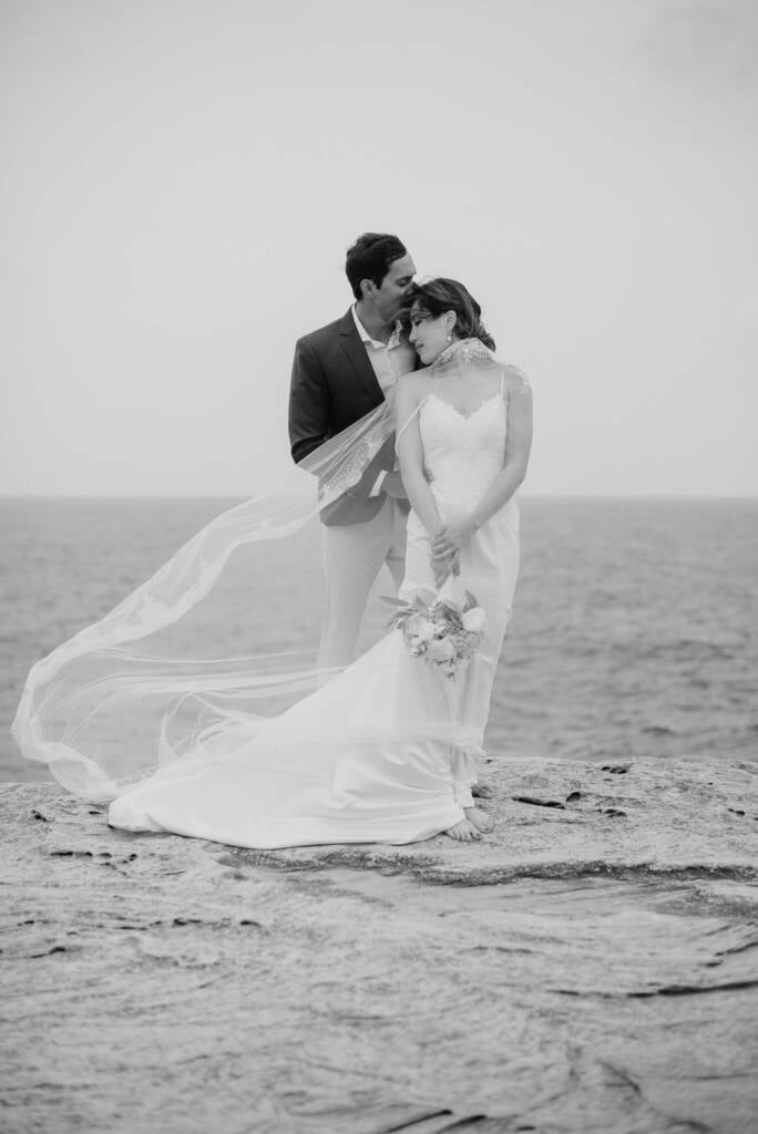Black-and-white portrait of Pierre kissing Jeannette’s forehead on the Cape Banks rock platform as her veil blows sideways.