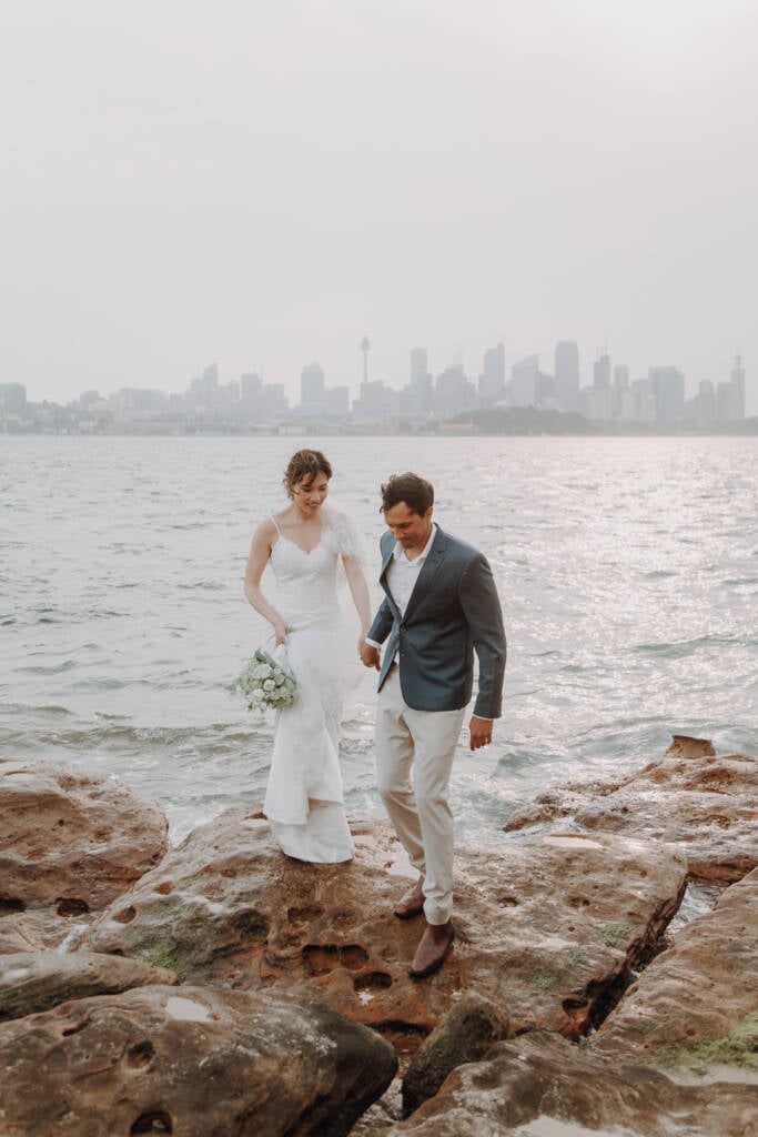 Jeannette and Pierre step across rocks at Bradleys Head with the Sydney skyline across the harbour.
