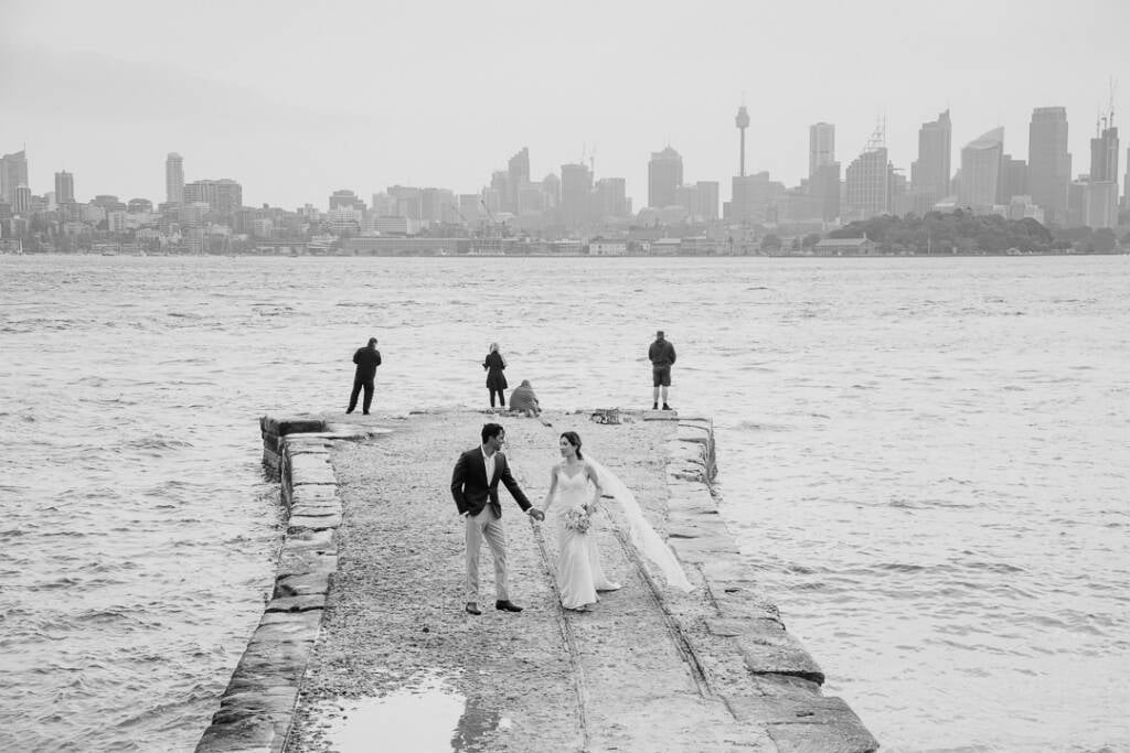 Black-and-white photo of Jeannette and Pierre holding hands on the Bradleys Head stone jetty, city skyline beyond.
