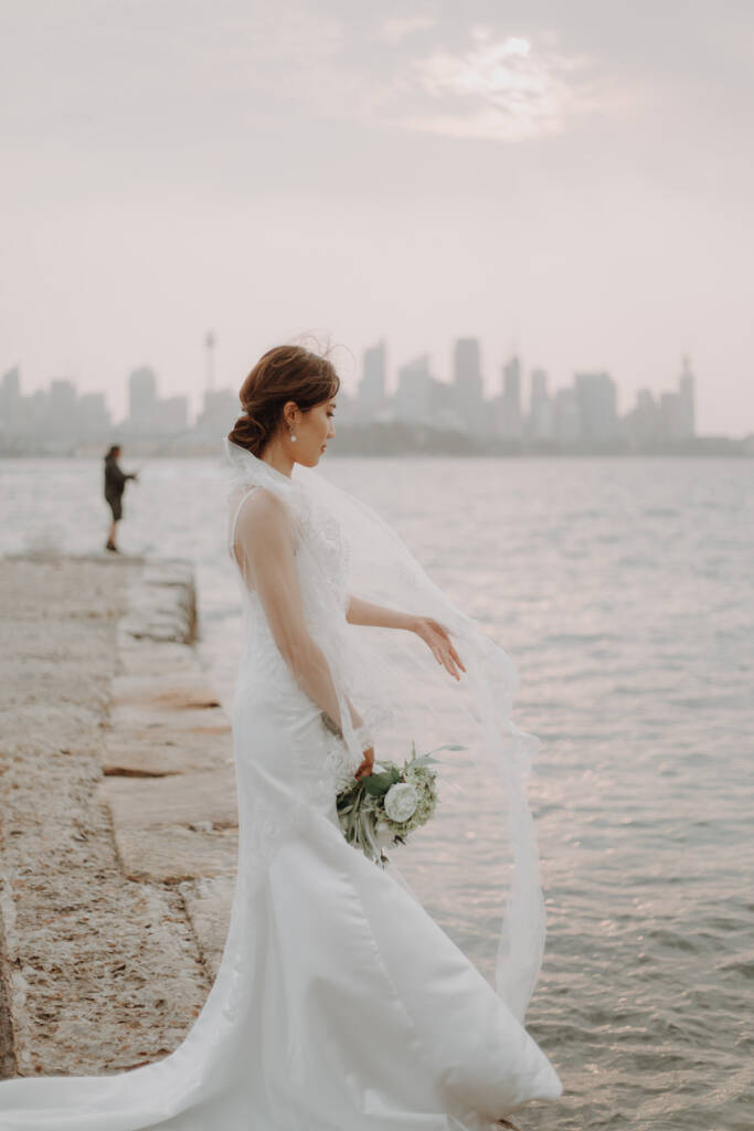 Jeannette stands in profile on the Bradleys Head jetty with her bouquet and veil beside the harbour.