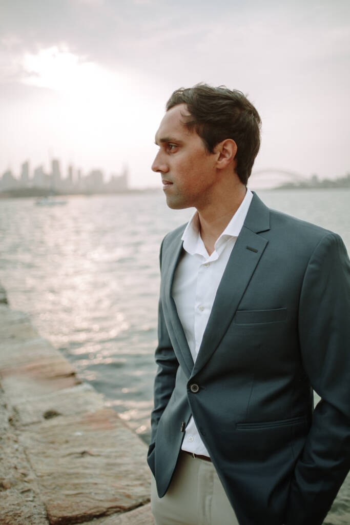 Portrait of Pierre beside the water at Bradleys Head, with the Sydney skyline and Harbour Bridge in soft focus.