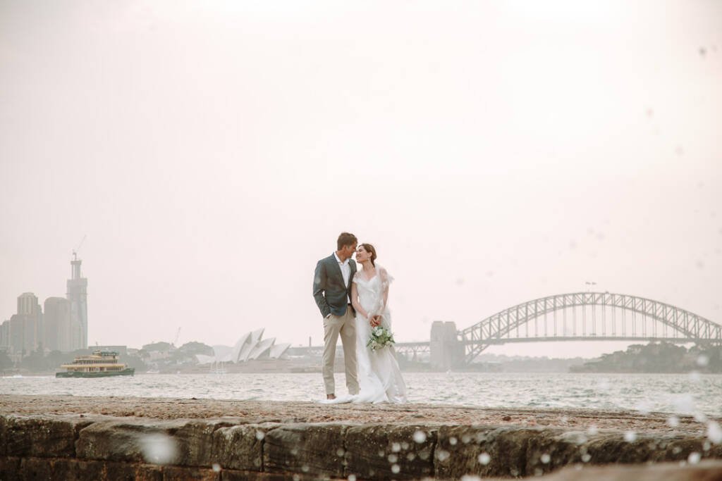 Jeannette and Pierre stand together on a stone sea wall at Bradleys Head, with the Sydney Opera House and Harbour Bridge behind.