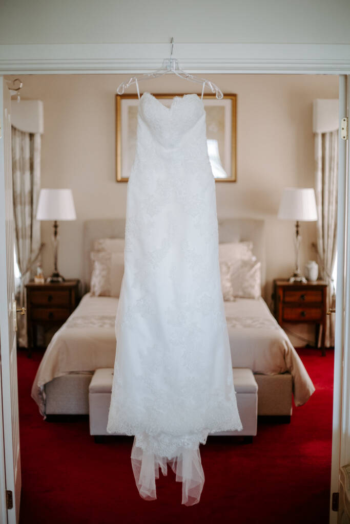 The strapless lace gown hangs in the doorway of a red carpeted bedroom with lamps either side.