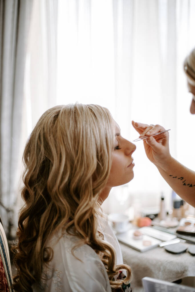 A steady hand shapes the bride's brow with a brush at the bright curtained window.