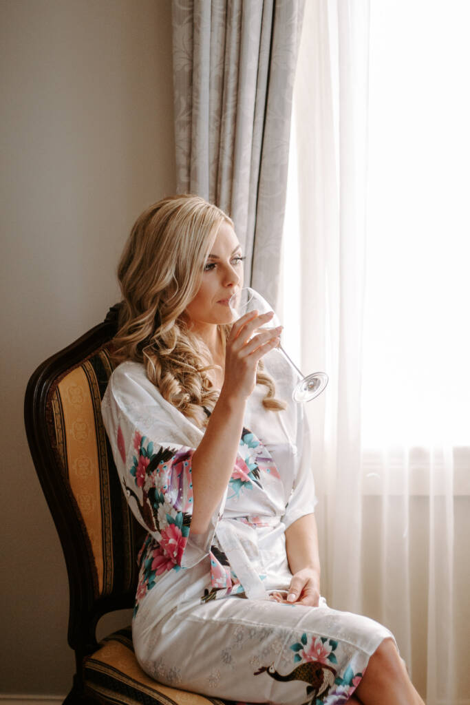 The bride takes a quiet sip of champagne in her crane print robe by the window.