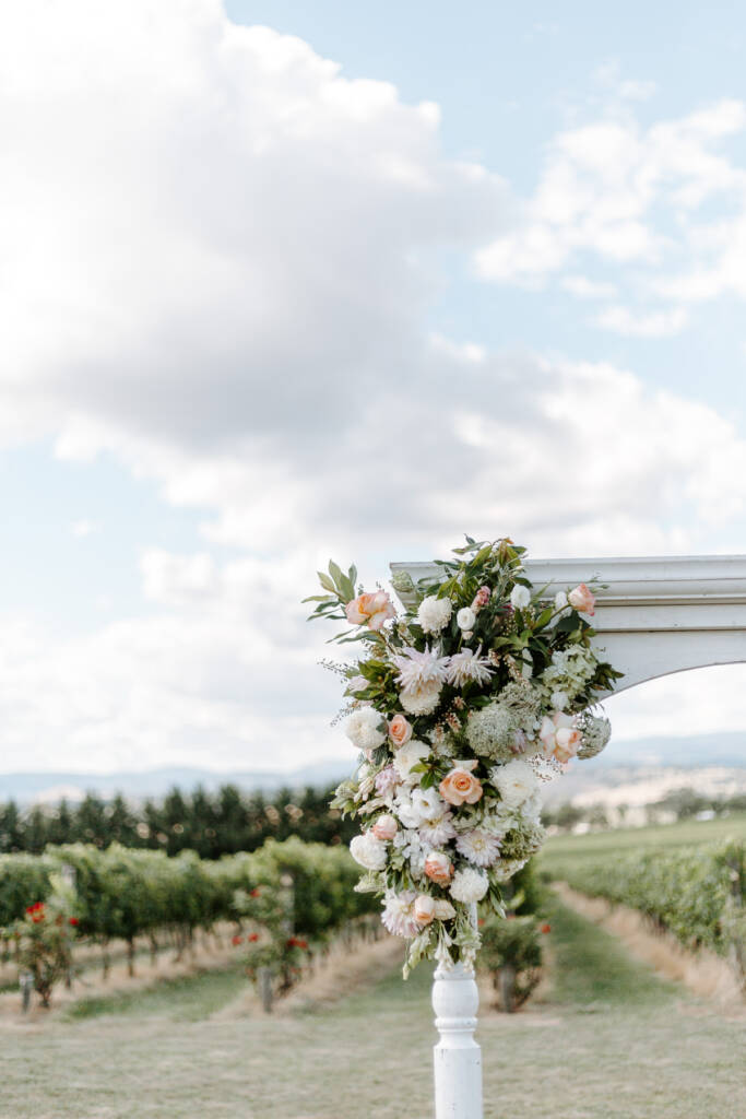 Blush flowers cover Tokar Estate's white ceremony arbour with vine rows beyond.