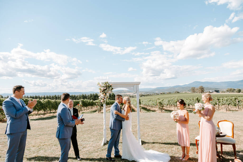 Danni and Jarrod share their first kiss as the wedding party applauds.