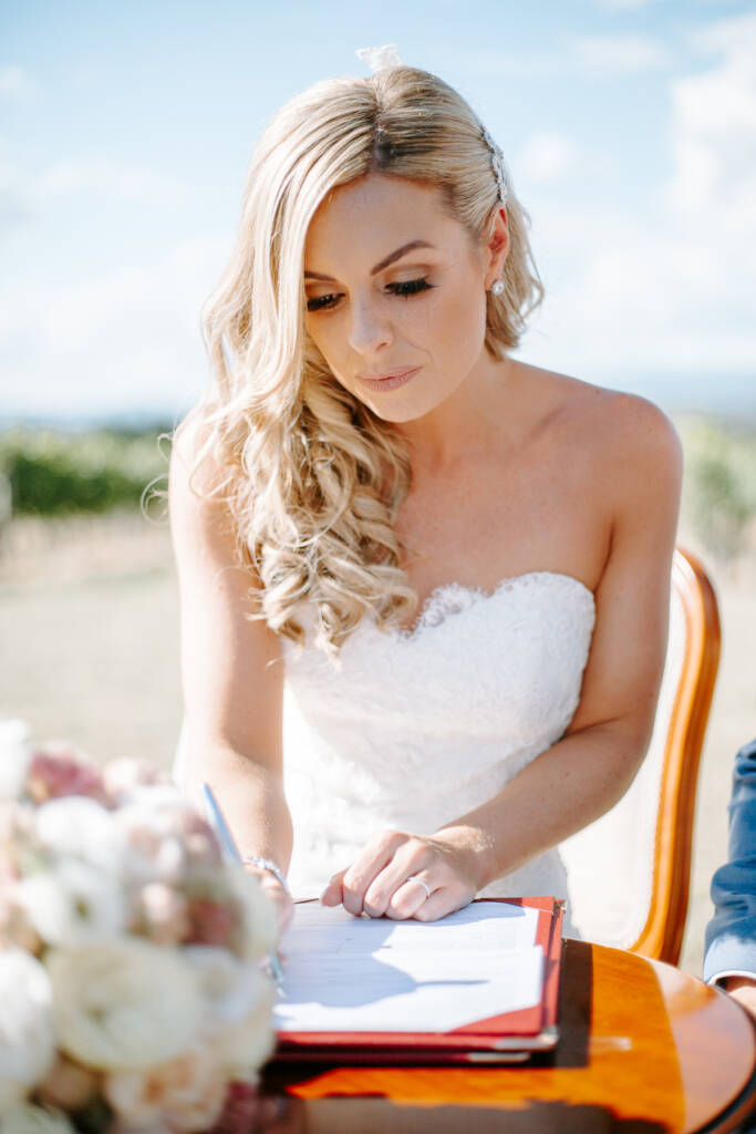 The bride bends over the register with the pen, curls falling across her shoulder.