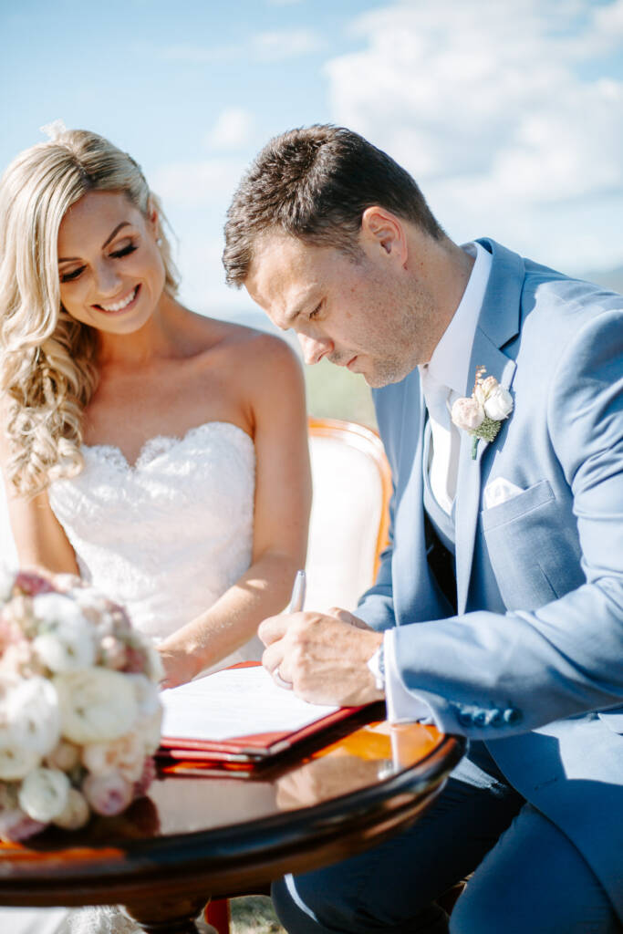 The groom's turn with the pen while the bride smiles at the page beside him.
