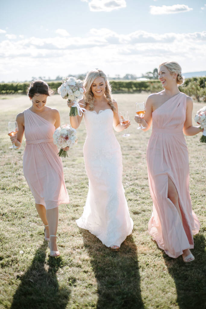 Danni and two bridesmaids walk together carrying bouquets and wine glasses.