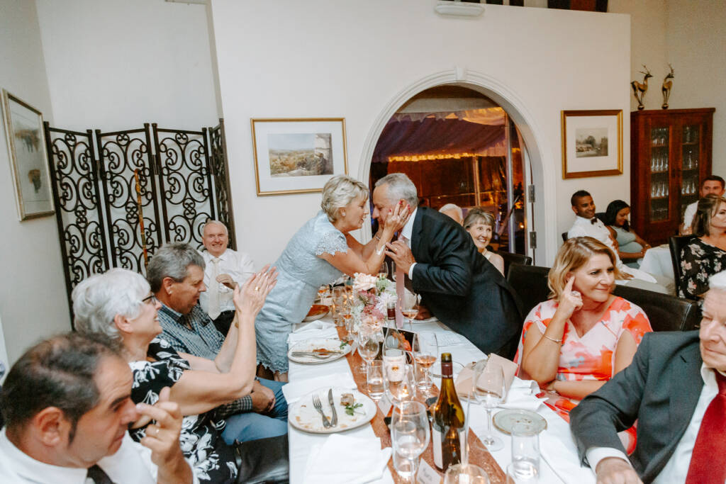 The woman in powder blue lace holds the face of the man in the navy suit, guests clapping.