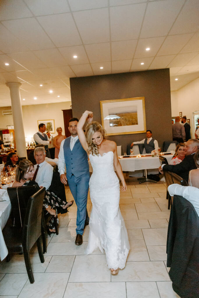 Jarrod leads Danni between reception tables with one hand raised.