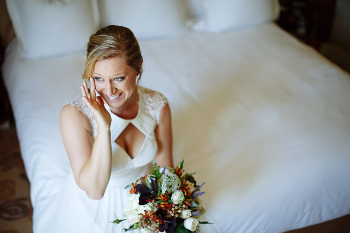 Lindsey presses away a happy tear on the white hotel bed, the bouquet in her lap.