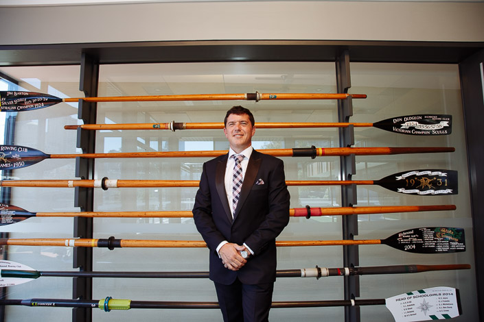 Checked tie and navy suit before a wall of champion oars