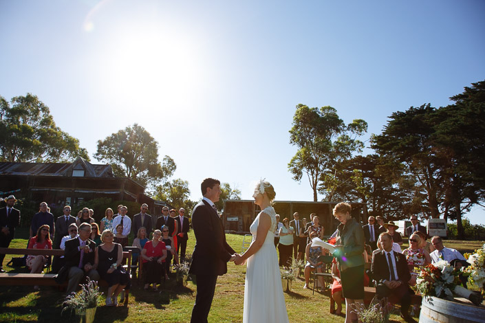Vows before the old farm sheds as the summer sun flares over the guests.