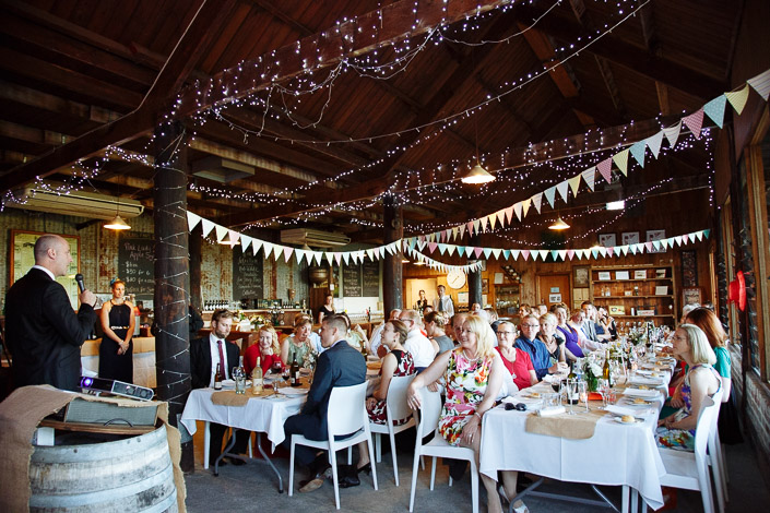 A man in a dark suit opens the night from a wine barrel lectern under the fairy lights.