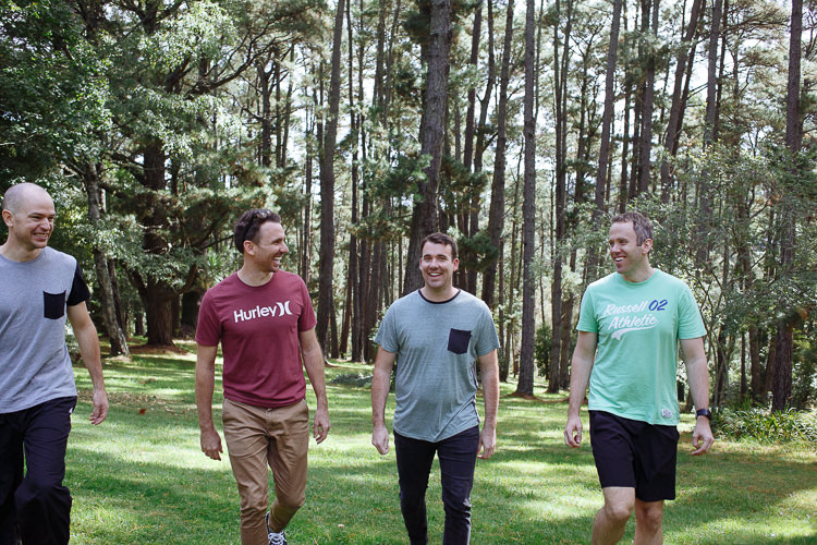 Russell and his groomsmen walk in casual clothes among tall trees at Yester Grange