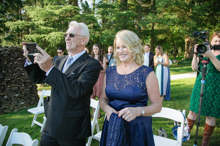 An older man takes a phone photo beside a smiling woman during the wedding