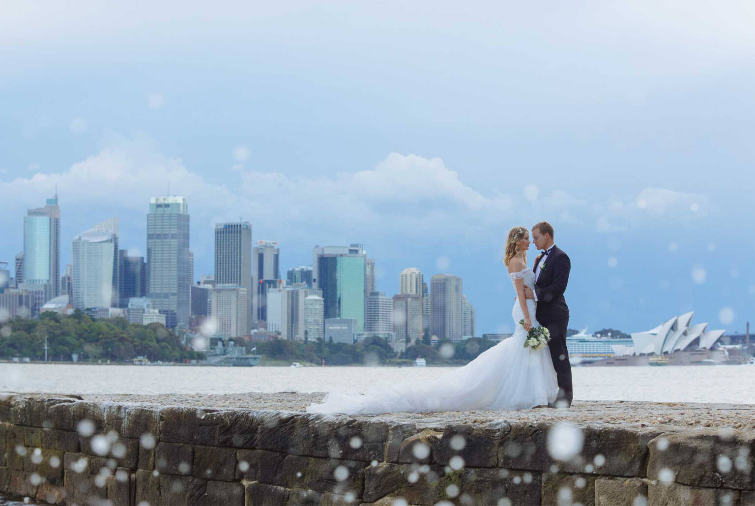 Across the harbour from the Sydney skyline, a couple on the sandstone sea wall as rain drifts through