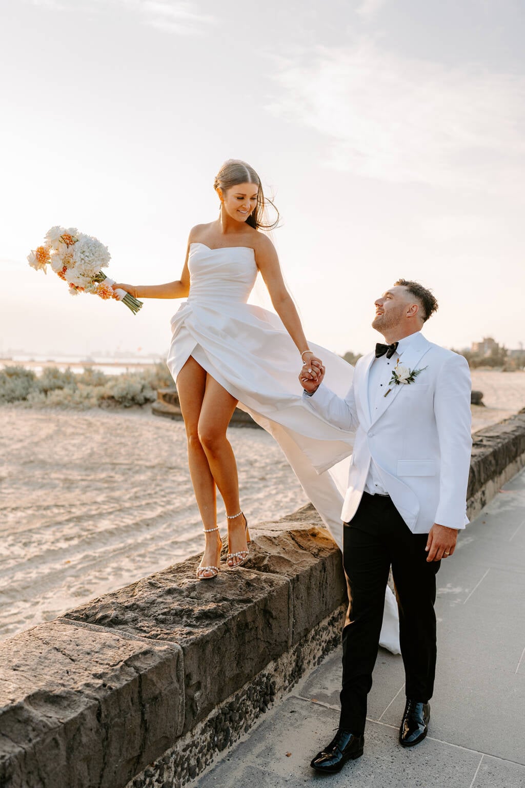 The bride on the bluestone wall at golden hour, her groom holding her hand — Harbour Room RMYS wedding, St Kilda