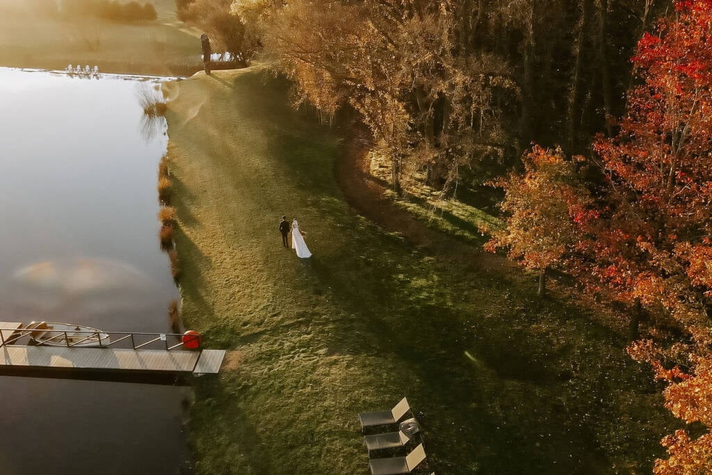 From above, Erica and Matthew cross the lakeside lawn in autumn light at Mona Farm, Braidwood