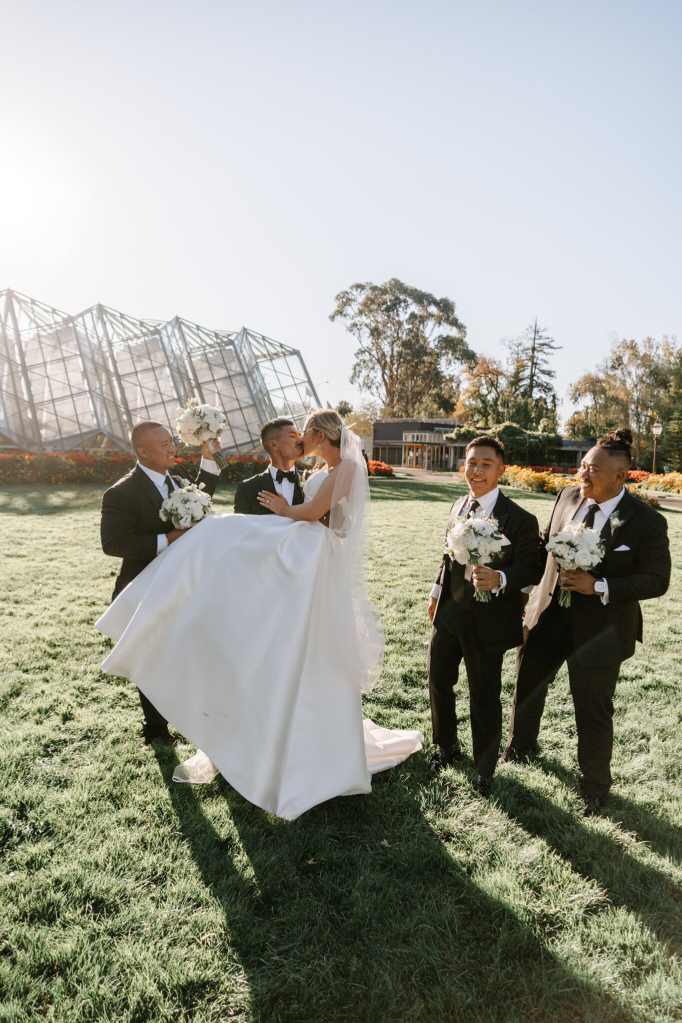 Johnny lifts Georgie as the groomsmen laugh beside the glasshouse at Ballarat Botanical Gardens.