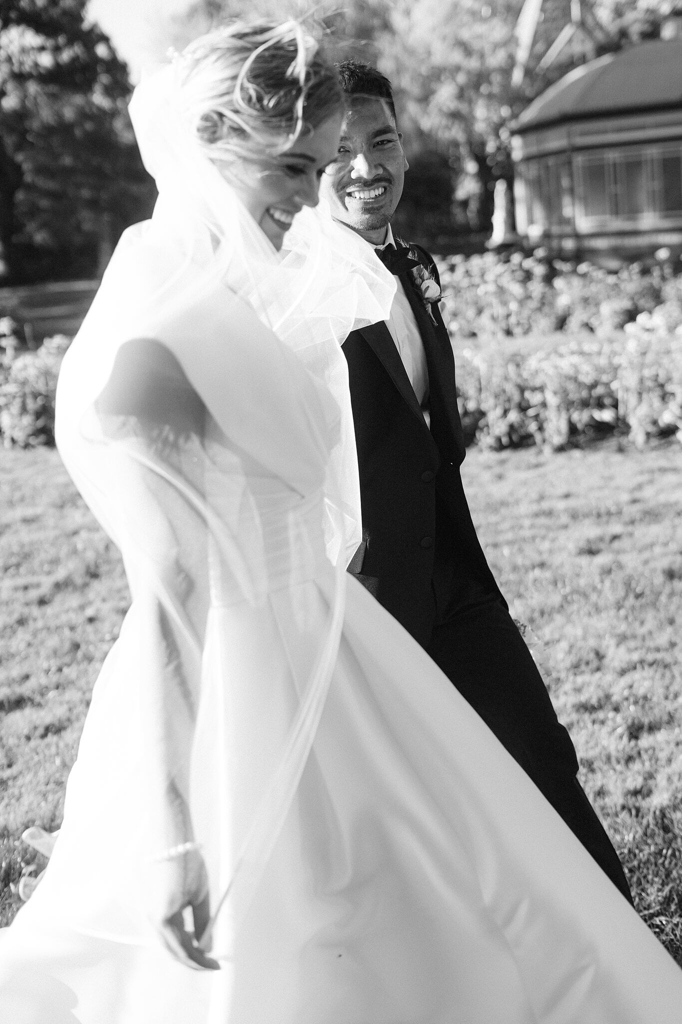 The wind throws Georgie’s veil across her face as Johnny laughs in Ballarat Botanical Gardens.