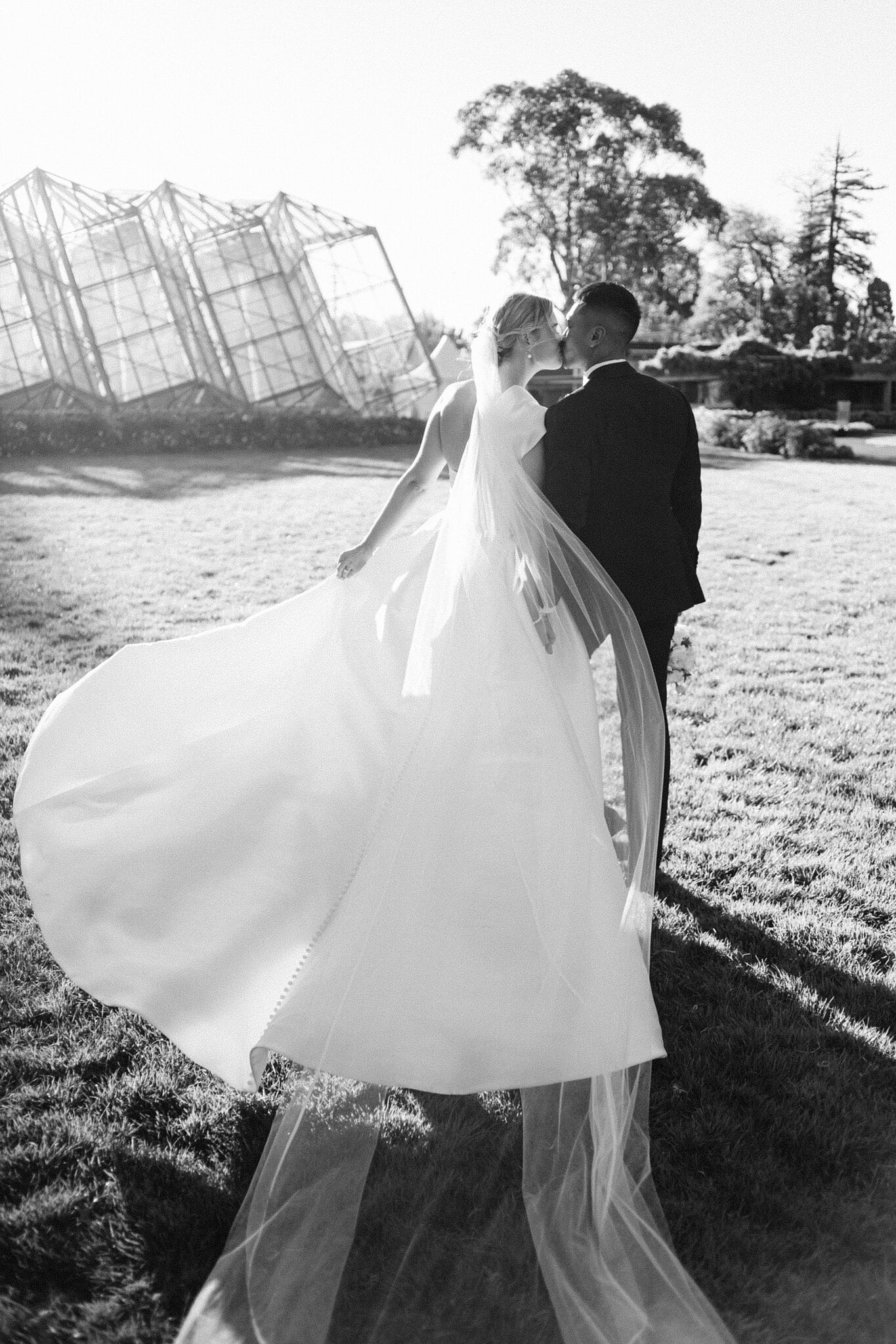Veil and gown caught by the wind as the bride and groom kiss beside the glasshouse at Ballarat Botanical Gardens