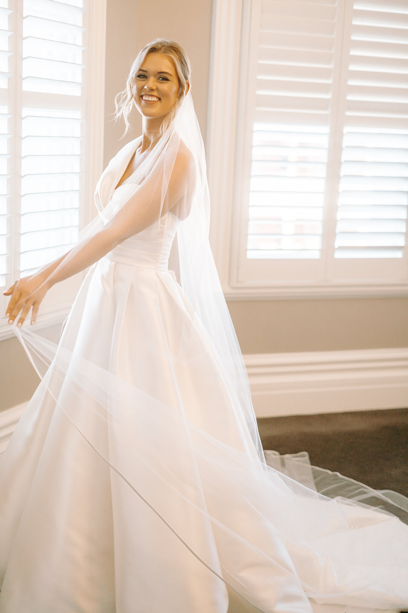 Georgie laughs as her veil drifts behind her, dressed and ready by the shuttered window.