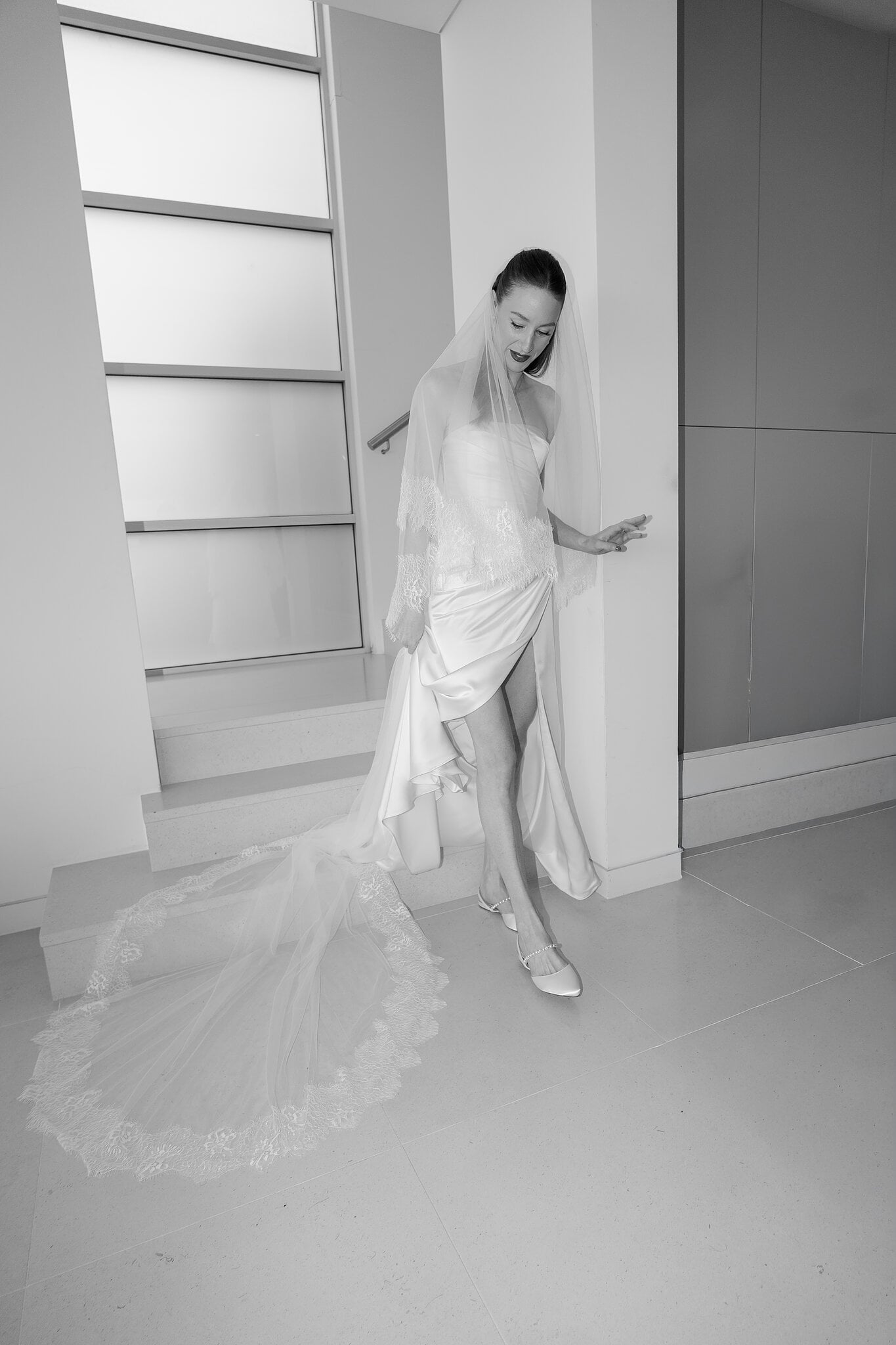 Michele steps down the stairs, her lace-trimmed veil pooling on the floor behind her.