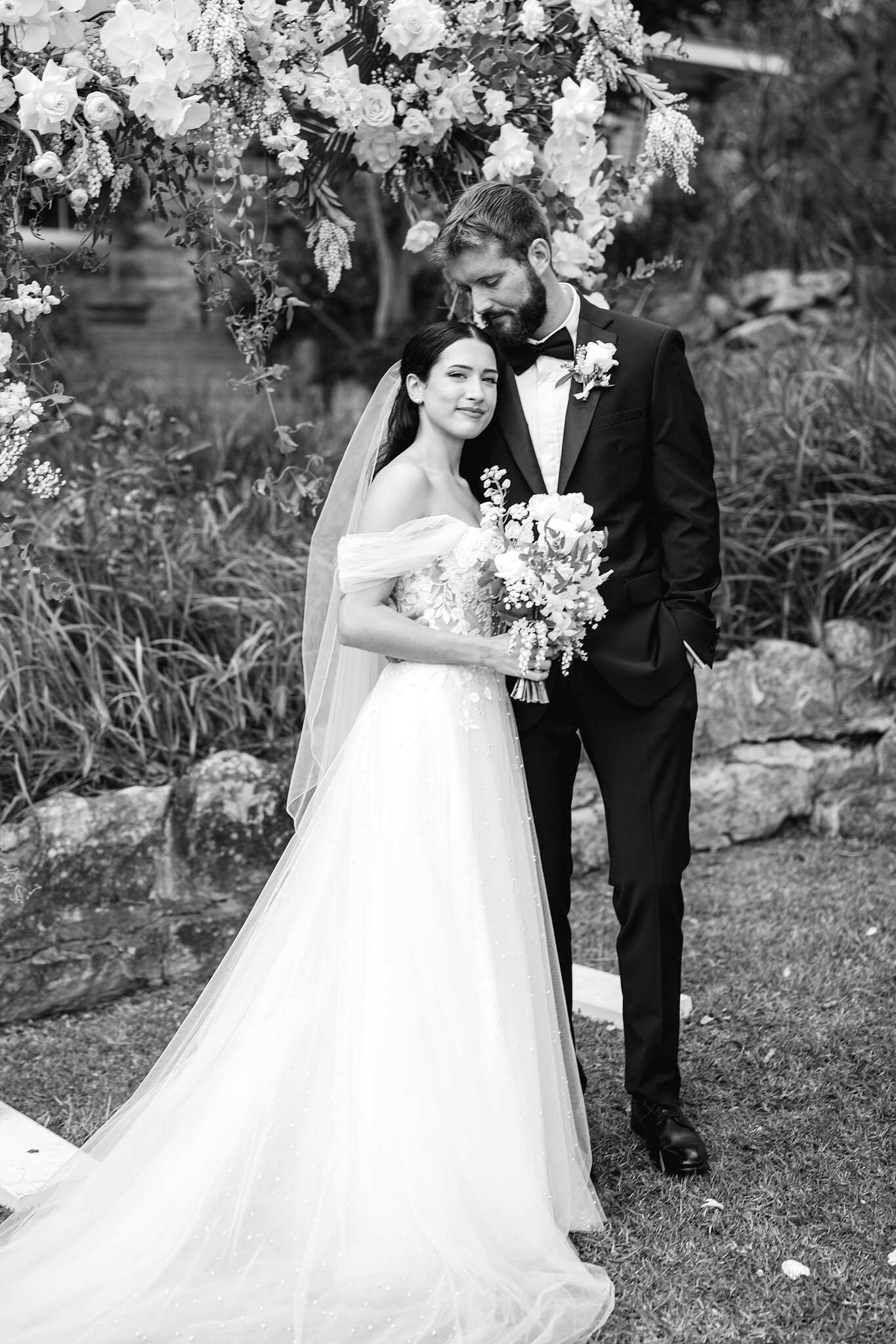 Alexa and Chris beneath the white floral arch on the ceremony lawn at Burnt Orange.