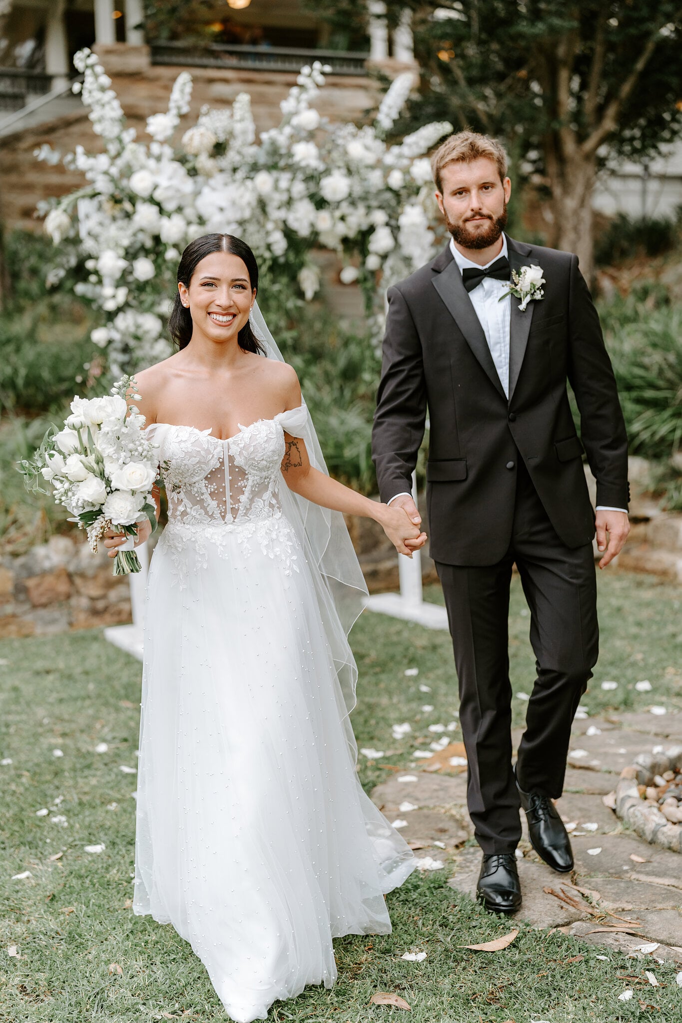 Alexa and Chris walk hand in hand past the white floral ceremony arbour at Burnt Orange.