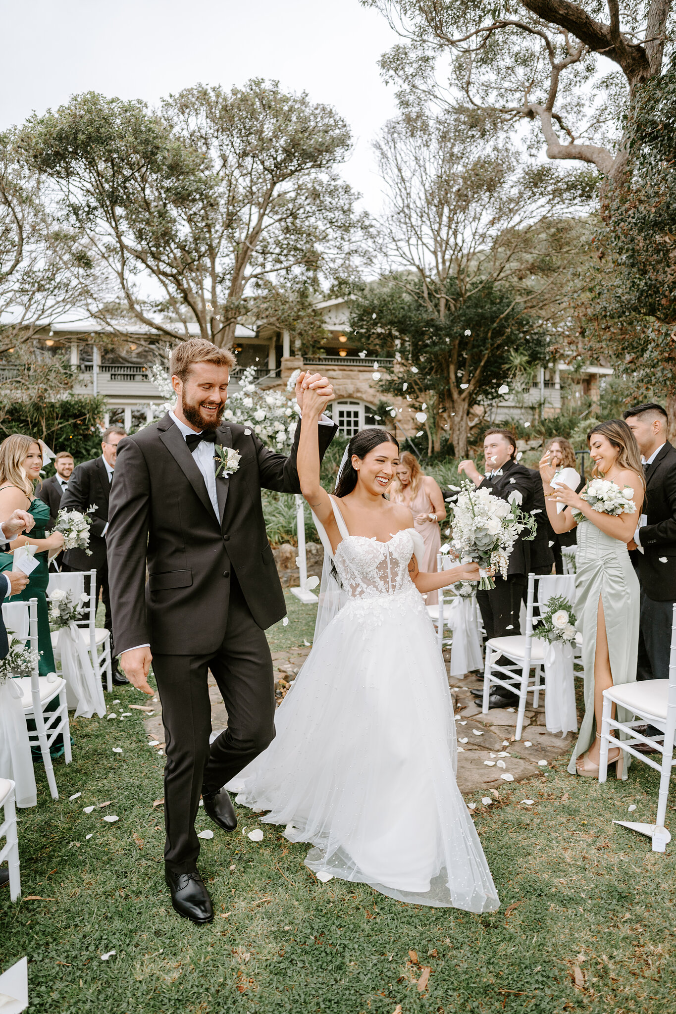 Just married, Alexa and Chris cheer back up the aisle through a shower of petals on the Burnt Orange lawn.