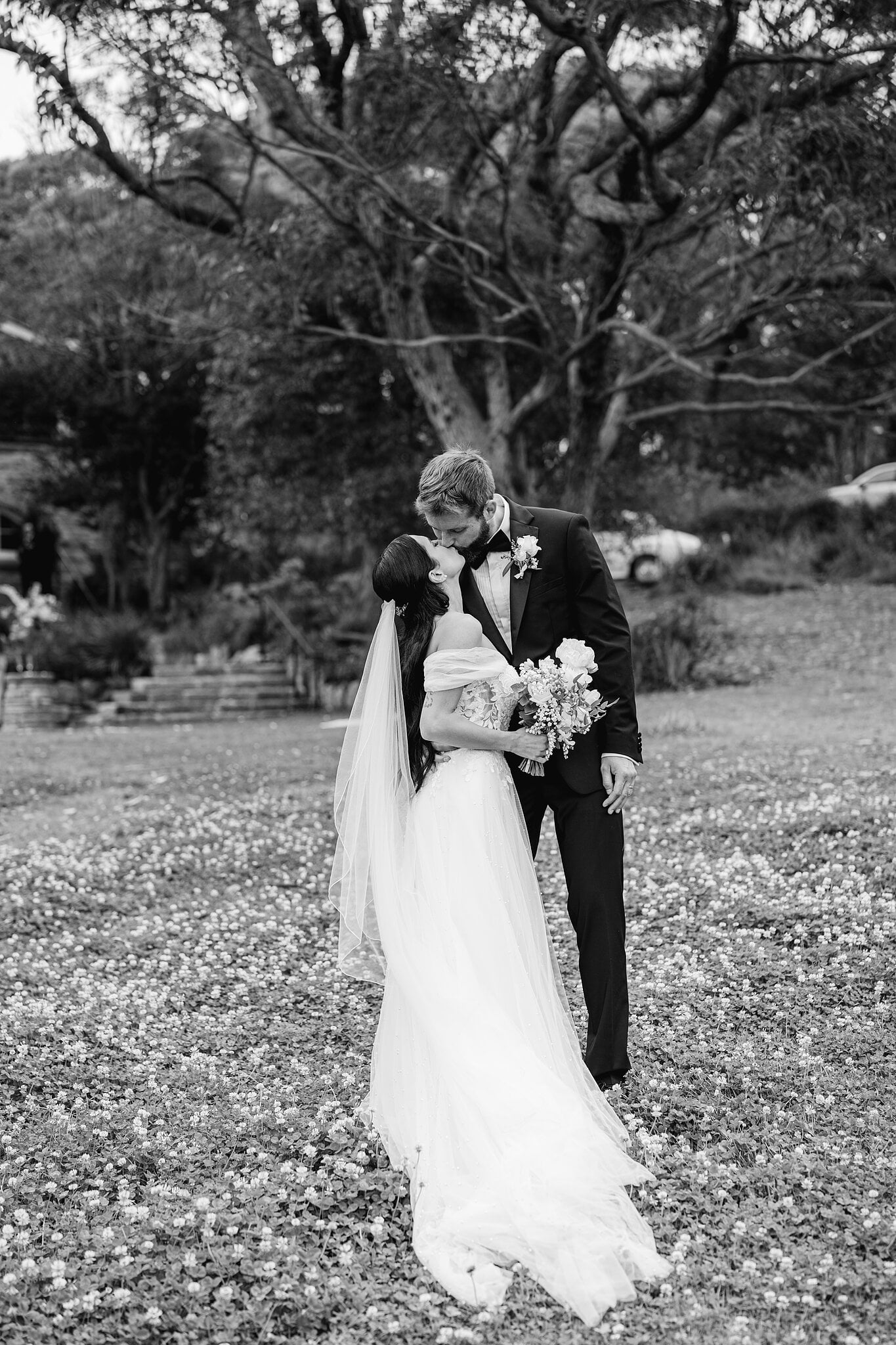 A kiss on the leaf-strewn lawn under a sprawling old tree at Burnt Orange, stone steps behind.