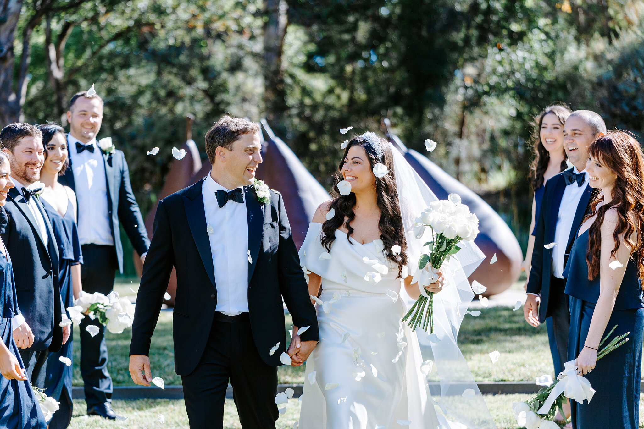 Helena and Xavier walk between wedding attendants as petals fall in a Canberra garden.