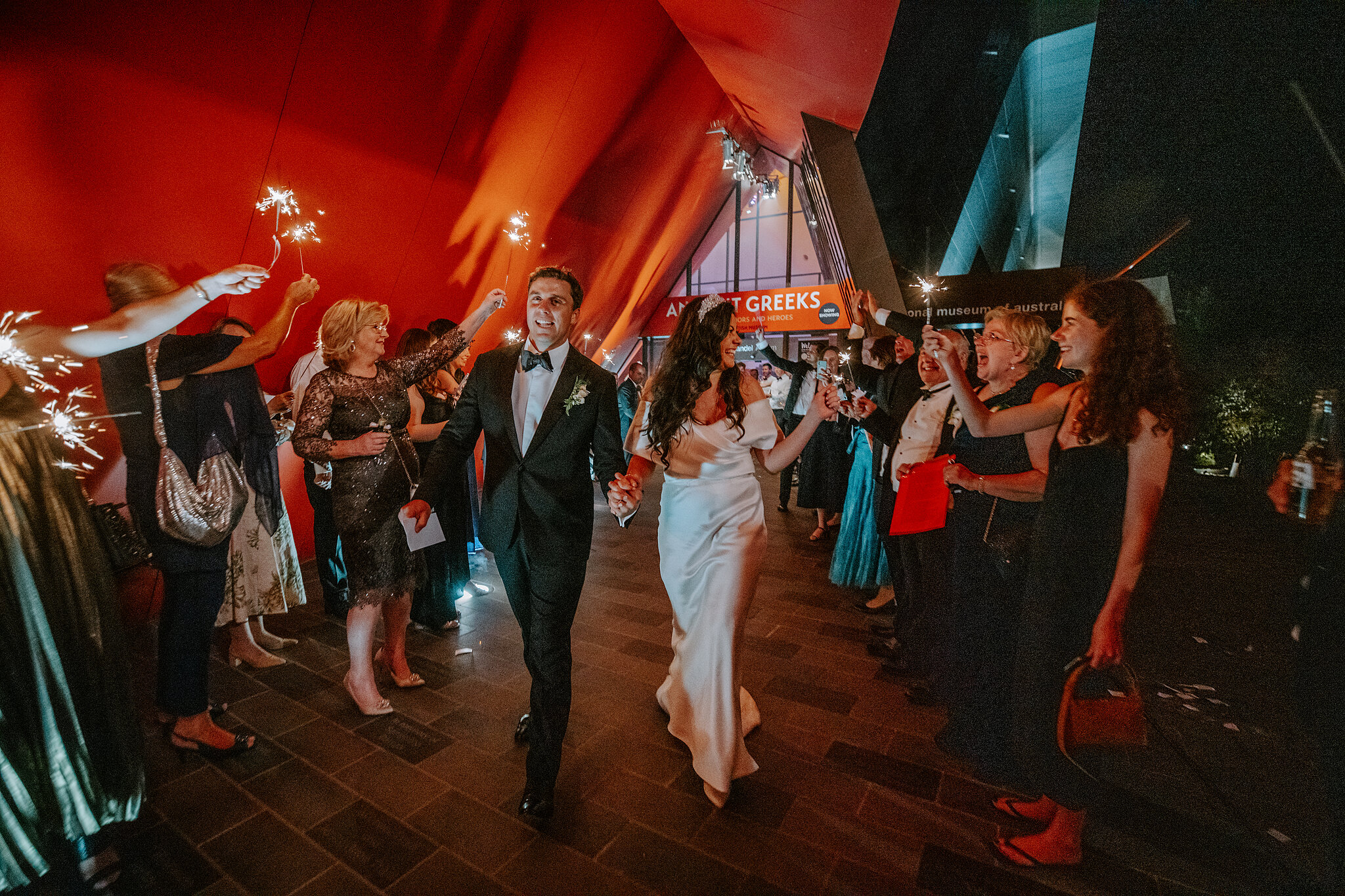 Helena and Xavier walk between guests holding sparklers outside the National Museum of Australia.