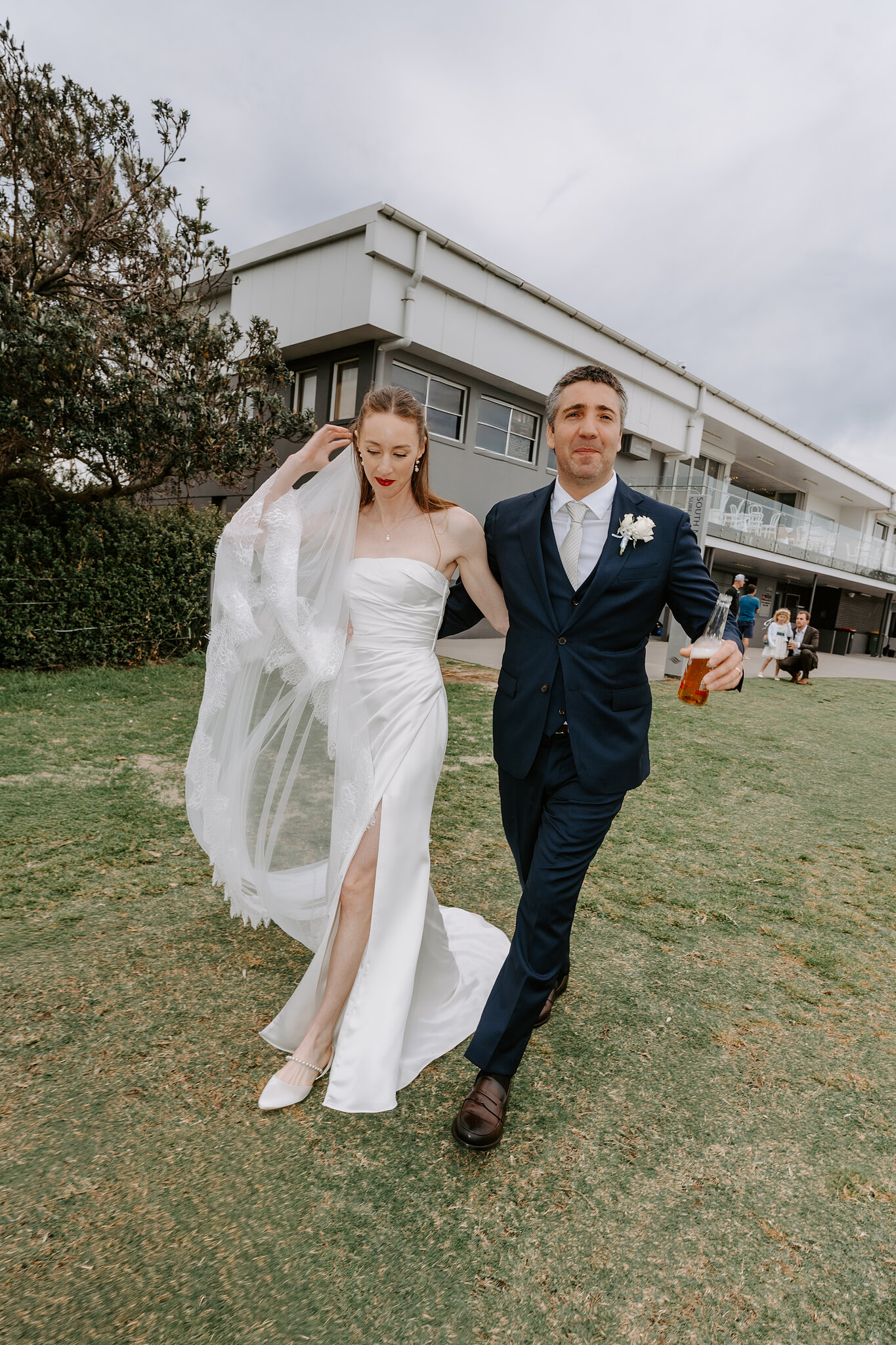 Michele and Nico stride across the lawn at Horizons South Maroubra, a beer in his hand and her veil in the wind.