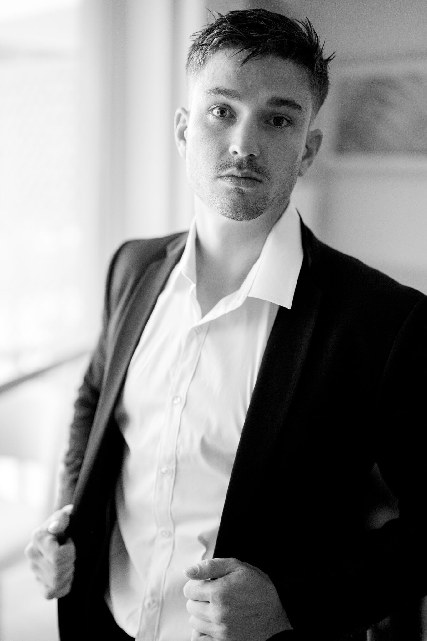 Lane shrugs on his black suit jacket in soft window light as he gets ready.