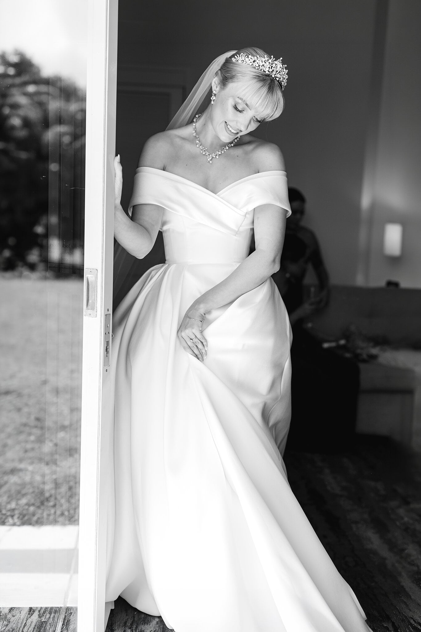 Ashley pauses at the doorway in her off-the-shoulder gown, her tiara catching the light.