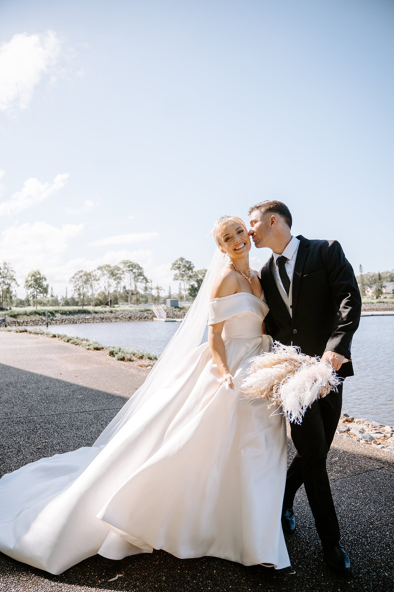 Lane kisses Ashley’s cheek by the water as she holds her pampas bouquet.