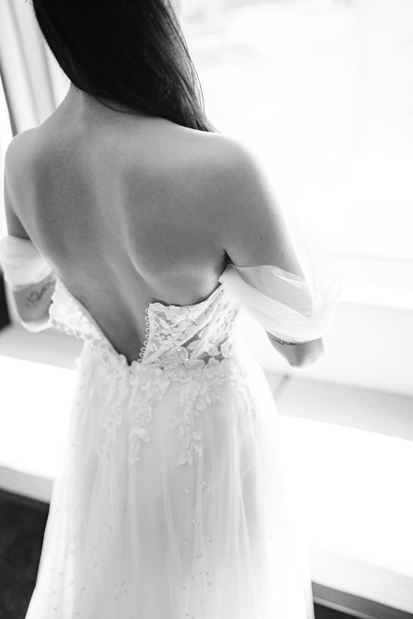 The lace corset and off-the-shoulder tulle sleeves across the back of Alexa’s gown, lit by the window.