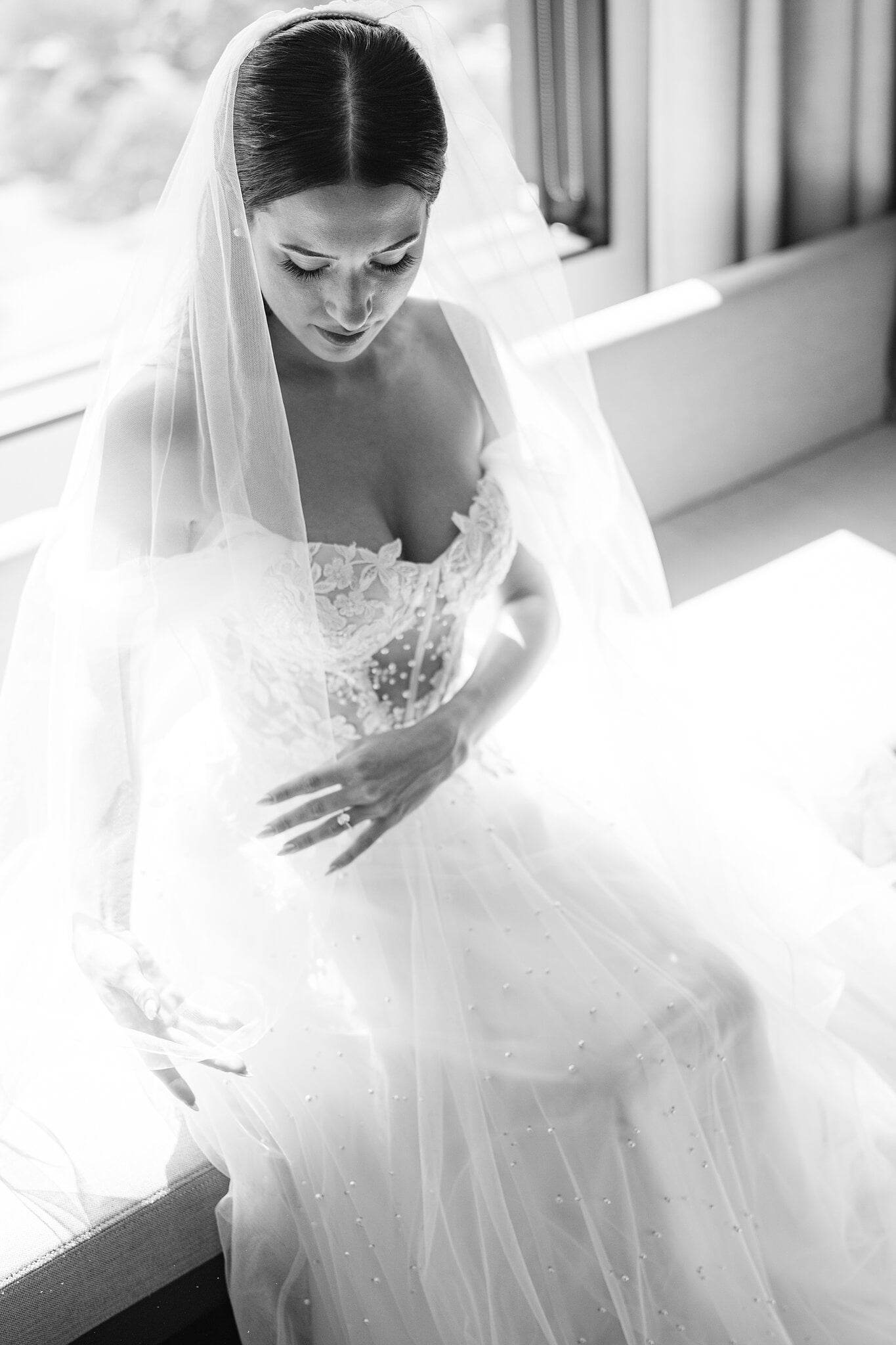 Veil on, Alexa smooths the pearl-scattered tulle of her gown beside the window.