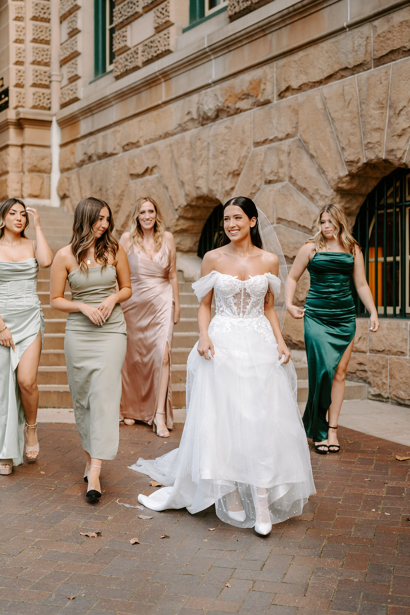 Alexa walks out with her bridesmaids in sage, blush and emerald satin past the sandstone of InterContinental Sydney.