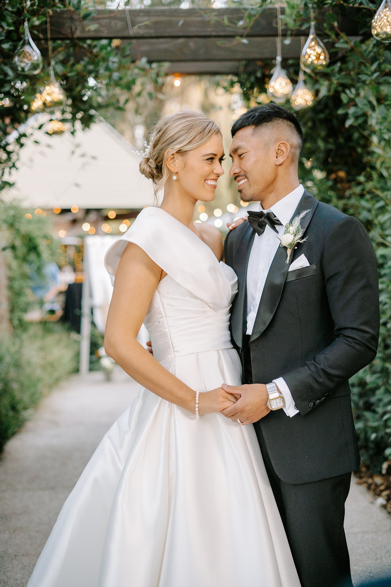 Georgie and Johnny share a quiet moment, foreheads close, as guests gather beyond the pergola.