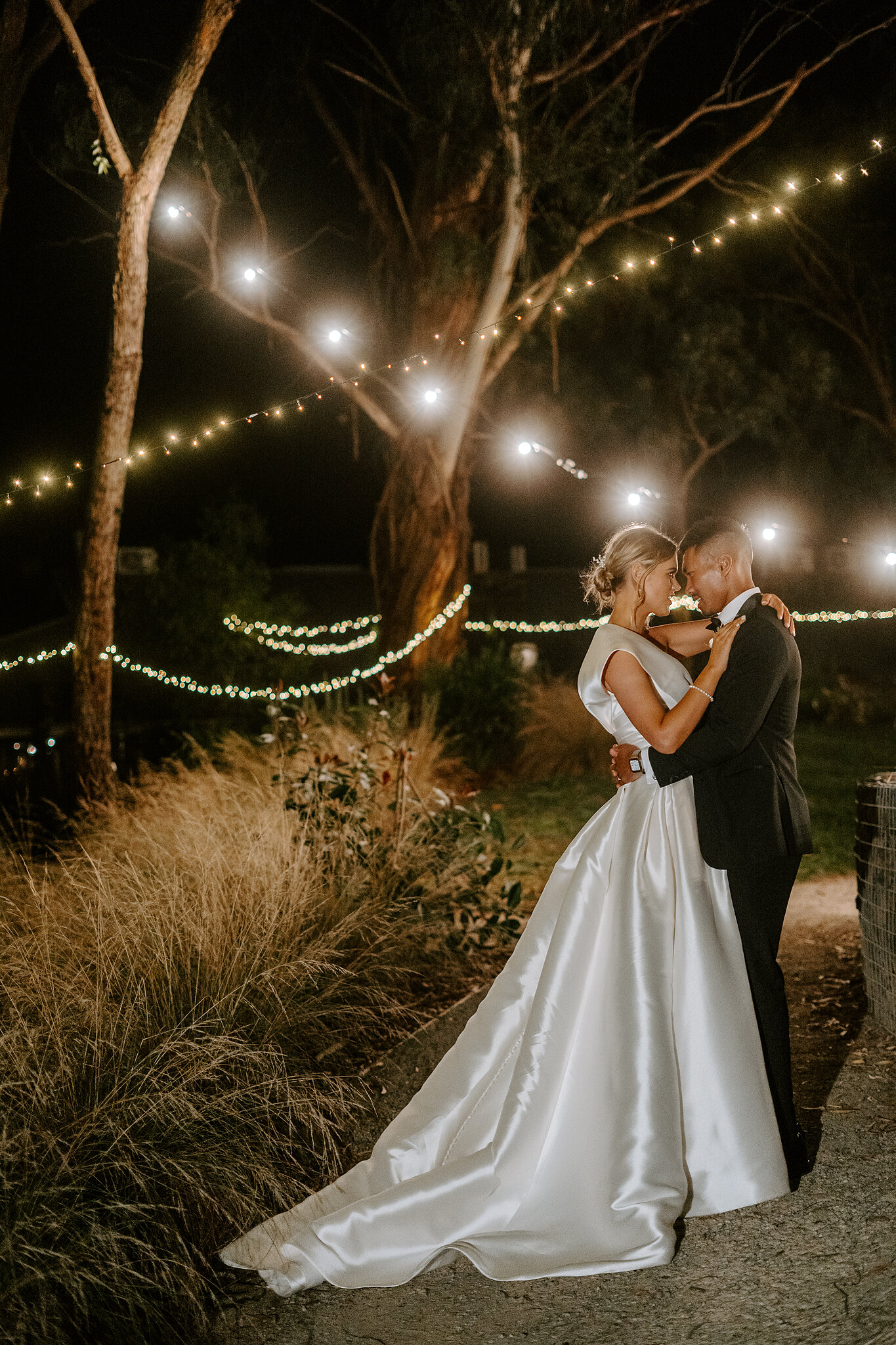 Georgie and Johnny embrace under festoon lights strung through the gum trees at Mercure Ballarat.